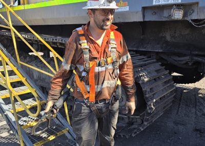 A man in an orange safety vest stands on a ladder beside a large truck, overseeing maintenance or loading tasks.